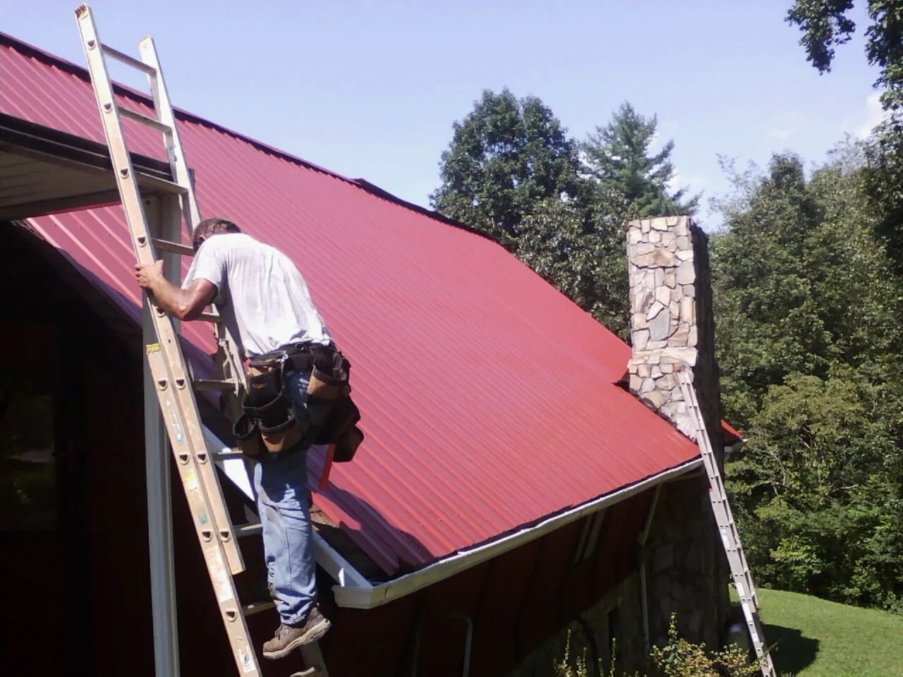 Roofer on a ladder beside a red metal roof and stone chimney — Soffit & Fascia Repair by Roofing Brothers in Morrilton